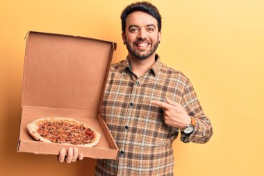İspanyol asıllı genç adam pizza kutusunu tutuyor. Gülümsüyor, mutlu bir şekilde parmakla işaret ediyor. 