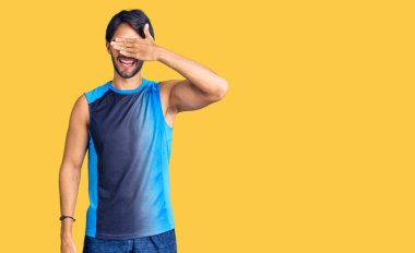 Spor kıyafetleri giyen yakışıklı İspanyol adam. Gülümsüyor ve elleri yüzünde, şaşırtmak için gözlerini kapatıyor. kör kavram. 