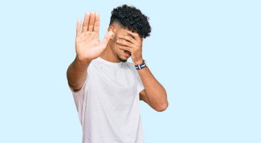 Gündelik beyaz tişört giyen genç Arap adam elleriyle gözlerini kapatıyor ve üzgün ve korku ifadeleriyle jest yapıyor. utangaç ve olumsuz kavram. 