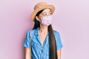 Yazın tıbbi maske takan genç Çinli bir kadın, yan tarafa bakıyor, rahat bir görünüm takınıyor ve kendine güvenen bir gülümseme takınıyor.. 