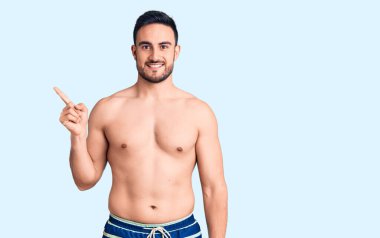 Yüzünde kocaman bir gülümseme olan mayo giyen genç ve yakışıklı bir adam, kameraya el ile işaret ediyor.. 