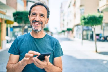 Sakallı orta yaşlı bir adam dışarıda mutlu bir şekilde gülümsüyor akıllı telefon kullanıyor.