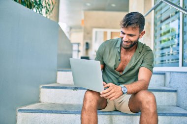 Merdivenlerde dizüstü bilgisayar kullanarak gülümseyen İspanyol genç adam.