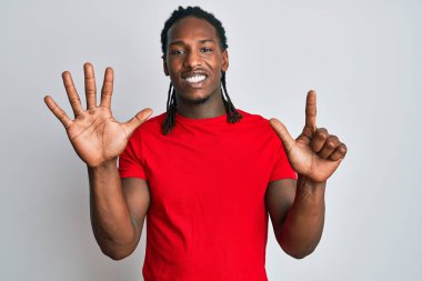Saçları örgülü Afro-Amerikalı bir adam 7 numarayı gösterip gösterirken kendinden emin ve mutlu bir şekilde gülümsüyor.. 