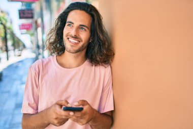 Duvara dayanmış akıllı telefon kullanan, mutlu gülümseyen genç İspanyol adam.