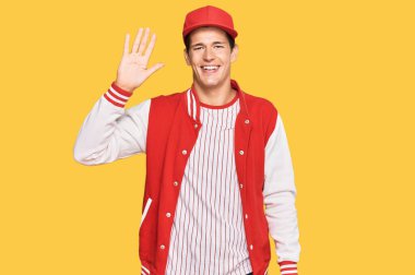 Beyzbol üniforması giyen yakışıklı beyaz adam beş numarayı gösterip işaret ederken kendinden emin ve mutlu bir şekilde gülümsüyor.. 