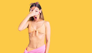 Güzel esmer, bikini giyen ve güneş gözlüklü bir kadın. Şok içinde, yüzünü ve gözlerini kapayarak utangaç ifadesiyle parmaklarından bakıyor.. 