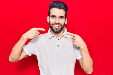 Sakallı, sıradan polo giyen neşeli gülümseyen ve parmaklarıyla ve ağzıyla işaret eden yakışıklı bir genç. diş sağlığı kavramı. 