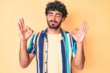 Kıvırcık saçlı, ayı kılıklı, yaz gömleği giyen, güneş gözlüklü, gözleri kapalı, meditasyon yaparken parmaklarıyla gülümseyen yakışıklı bir genç. yoga konsepti. 