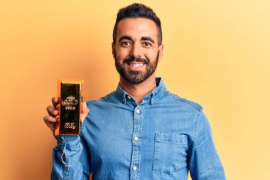 Altın külçeli İspanyol genç adam pozitif ve mutlu görünüyor. Duruyor ve gülümsüyor. Kendine güvenen bir gülümsemeyle dişlerini gösteriyor. 