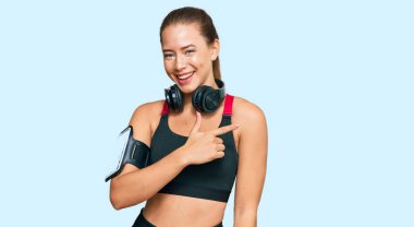 Güzel sarışın kadın spor kıyafetleri giyiyor ve kulaklık takıyor. Yüzünde gülücükler, el ve parmaklarıyla mutlu ve doğal bir ifade var. 