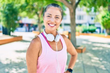 Orta yaşlı sporcu kadın parkta kulaklık takarak mutlu gülümsüyor.