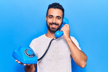 İspanyol asıllı genç adam elinde kaliteli bir telefon tutuyor. Pozitif ve mutlu görünüyor. Duruyor ve gülümsüyor. 