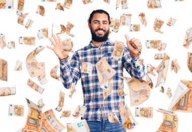 Genç Arap adam gündelik giysiler giyer ve altı numaralı parmaklarıyla işaret ederken kendinden emin ve mutlu bir şekilde gülümser..