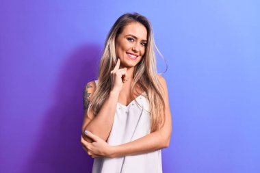 İzole edilmiş mor arka planda duran beyaz tişört giyen genç ve güzel sarışın kadın kameraya kollarını kavuşturmuş ve çenesine dokunmuş şekilde gülümsüyor. Olumlu düşünüyorum..