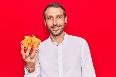 Elinde patates cipsi tutan genç yakışıklı adam pozitif ve mutlu görünüyor ve kendine güvenen bir gülümsemeyle dişlerini gösteriyor. 