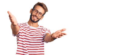 Sarışın, sakallı, sıradan kıyafetler giyen ve gözlüklü yakışıklı bir adam kameraya bakıp sarılmak için gülümsüyor. Mutluluğu kucaklayan neşeli bir ifade.. 