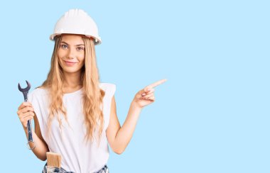 Beautiful caucasian woman with blonde hair wearing hardhat and builder clothes smiling happy pointing with hand and finger to the side  clipart