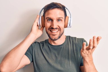 Genç yakışıklı adam kulaklık takarak müzik dinliyor. İzole edilmiş beyaz arka plan üzerinden başarıyı kutluyor. Gülümseyen bir gülümseme ve elini kaldırarak kazanan bir ifade.