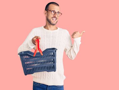 Süpermarket alışveriş sepetini tutan genç yakışıklı adam başparmağıyla yan tarafa bakıyor ve ağzı açık gülümsüyor. 