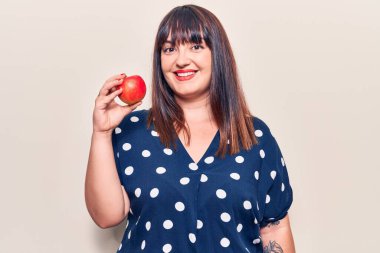 Elinde kırmızı elma tutan genç bir kadın. Pozitif ve mutlu görünüyor. Gülümsüyor ve kendine güvenen bir gülümsemeyle dişlerini gösteriyor. 