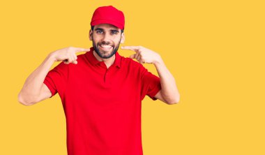 Sakallı, teslimat üniforması giyen, neşeli gülümseyen, parmakları ve ağzıyla işaret eden yakışıklı bir genç. diş sağlığı kavramı. 