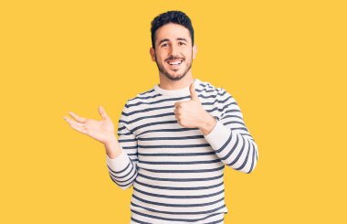 Latin asıllı genç adam, günlük kıyafetler giymiş, avuç içini gösteriyor ve baş parmaklarıyla iyi bir jest yapıyor, mutlu ve neşeli gülümsüyor. 