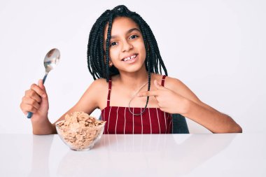 Masada oturmuş mısır gevreği yiyen, mutlu ve gururlu gülümseyen genç Afrikalı Amerikalı kız. 