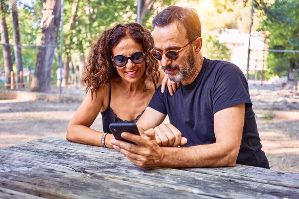 Medelålders Par Ler Glada Med Hjälp Smartphone Sitter Bänken Parken — Stockfoto