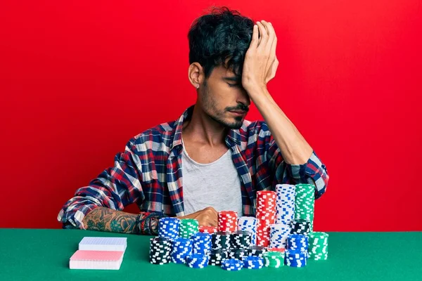 Young hispanic man sitting on the table with poker chips and cards surprised with hand on head for mistake, remember error. forgot, bad memory concept.