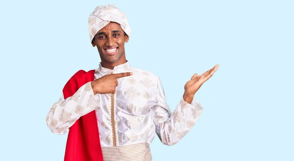 African handsome man wearing tradition sherwani saree clothes amazed and smiling to the camera while presenting with hand and pointing with finger.