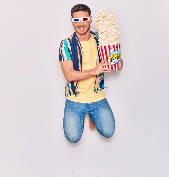 Young handsome hispanic man wearing 3d glasses smiling happy. Jumping with smile on face holding bucket of popcorn over isolated white background.