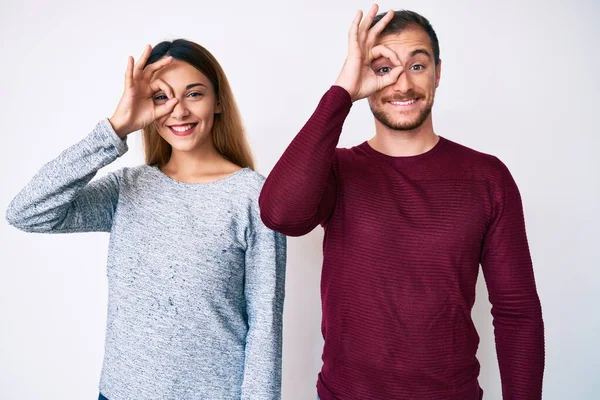 Beautiful couple wearing casual clothes smiling happy doing ok sign with hand on eye looking through fingers