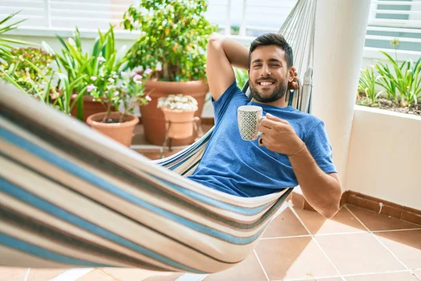 年轻的惊慌失措的男人在阳台上的吊床上放松地喝着一大杯咖啡 — 图库照片