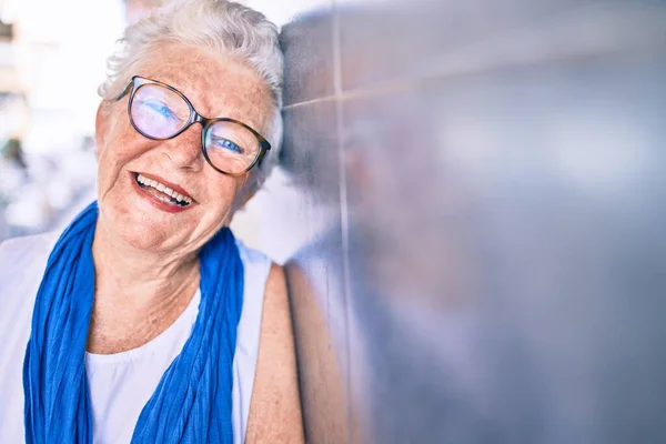 Elder senior woman with grey hair smiling happy leaning on the wall outdoors