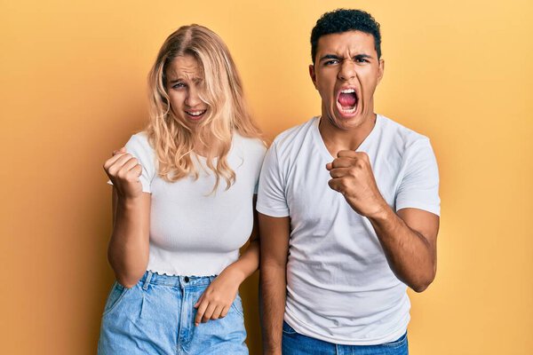 Молодая межрасовая пара в обычной белой футболке сердитая и безумная, поднимающая кулак, разочарованная и разъяренная, крича от злости. гнев и агрессивная концепция. 
