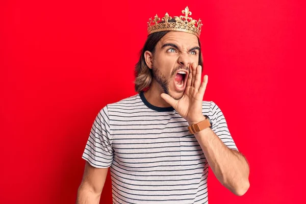 Young handsome man wearing prince crown shouting and screaming loud to side with hand on mouth. communication concept.
