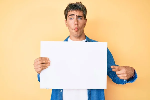 Young handsome man holding blank empty banner making fish face with mouth and squinting eyes, crazy and comical.