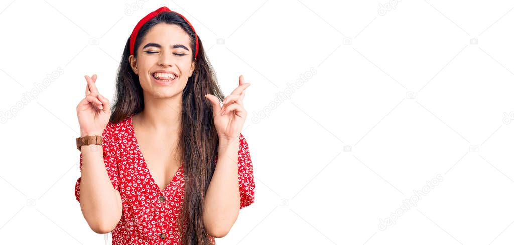 Brunette teenager girl wearing summer dress gesturing finger crossed smiling with hope and eyes closed. luck and superstitious concept. 