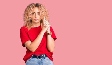 Kıvırcık saçlı, sıradan kırmızı tişörtlü, el hareketleriyle sembolik silah tutan, cinayet silahlarıyla oynayan, sinirli yüzlü genç sarışın kadın. 