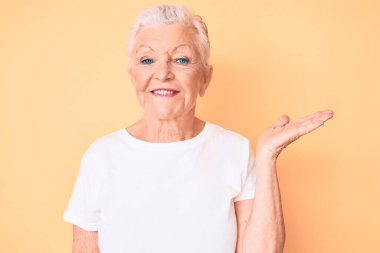 Son sınıf güzeli, mavi gözlü, gri saçlı, sarı arka planda klasik beyaz tişört giyen neşeli bir şekilde sunan ve kameraya bakan bir kadın.. 