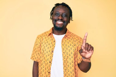 Saçları örgülü genç Afro-Amerikalı bir adam bir numarayı gösterip gösterirken kendinden emin ve mutlu bir şekilde gülümsüyor.. 