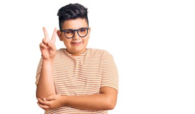 Günlük kıyafetler giyen ve gözlüklü küçük bir çocuk kameraya gülümseyen mutlu bir yüzle parmaklarıyla zafer işareti yapıyor. İki numara.. 