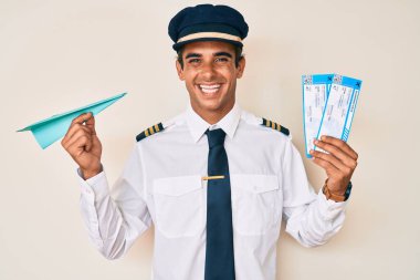 Uçak pilotu üniforması giyen genç İspanyol adam elinde kağıt uçak ve biniş kartı ile mutlu ve havalı bir gülümsemeyle gülümsüyor. diş gösterme. 