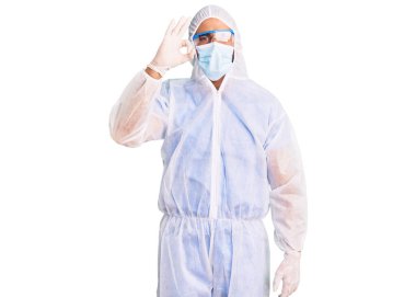 Doktor koruması, koronavirüs üniforması ve gülümseyen tıbbi maske takan genç İspanyol adam el ve parmaklarıyla imza atıyor. başarılı ifade. 