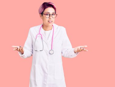 Pembe saçlı, doktor üniforması giyen, kafası karışmış ve kolları ve elleri havada genç ve güzel bir kadın. şüphe kavramı. 