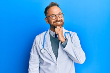 Doktor üniforması ve steteskop giyen yakışıklı bir adam. Kameraya kendinden emin bakıyor. Kolları çapraz ve elleri çenesinde. Olumlu düşünmek. 