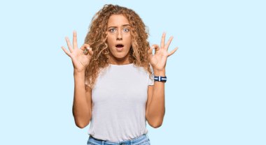 Beyaz günlük tişört giyen güzel beyaz kız şaşırmış ve şok olmuş bir şekilde parmaklarıyla onay sembolü yapıyor. çılgın ifade 