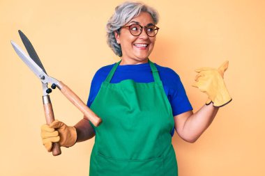 Bahçıvan önlüğü giymiş, elinde makas tutan, ağzı açık gülümseyen yaşlı İspanyol kadın. 