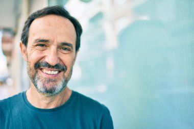 Orta yaşlı, sakallı, mutlu, açık havada duvara yaslanmış bir adam.